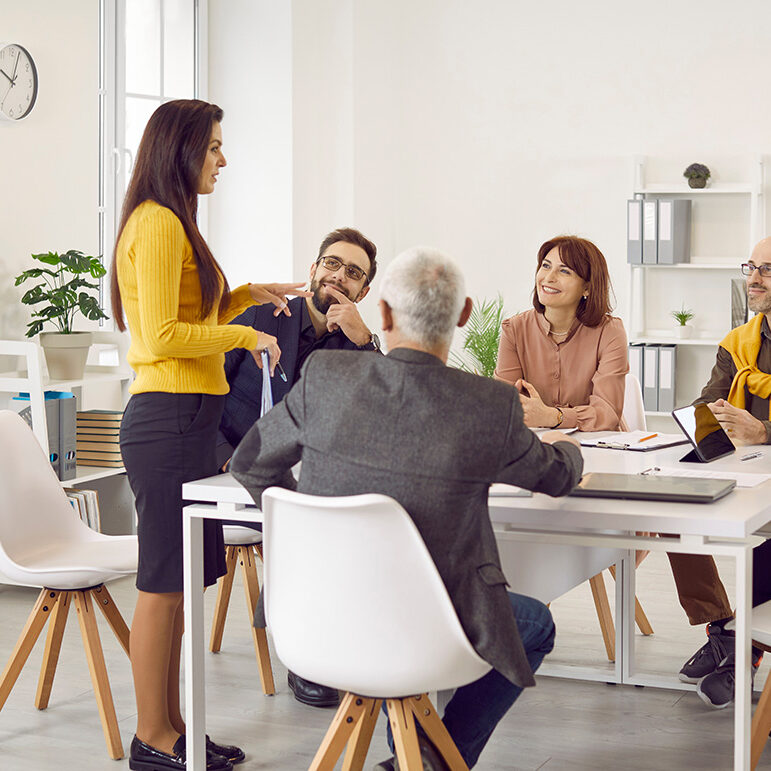 Diverse business team having a work meeting in a modern office. Group of happy multiethnic people sitting around a table with laptops and tablets and listening to a young woman making a suggestion