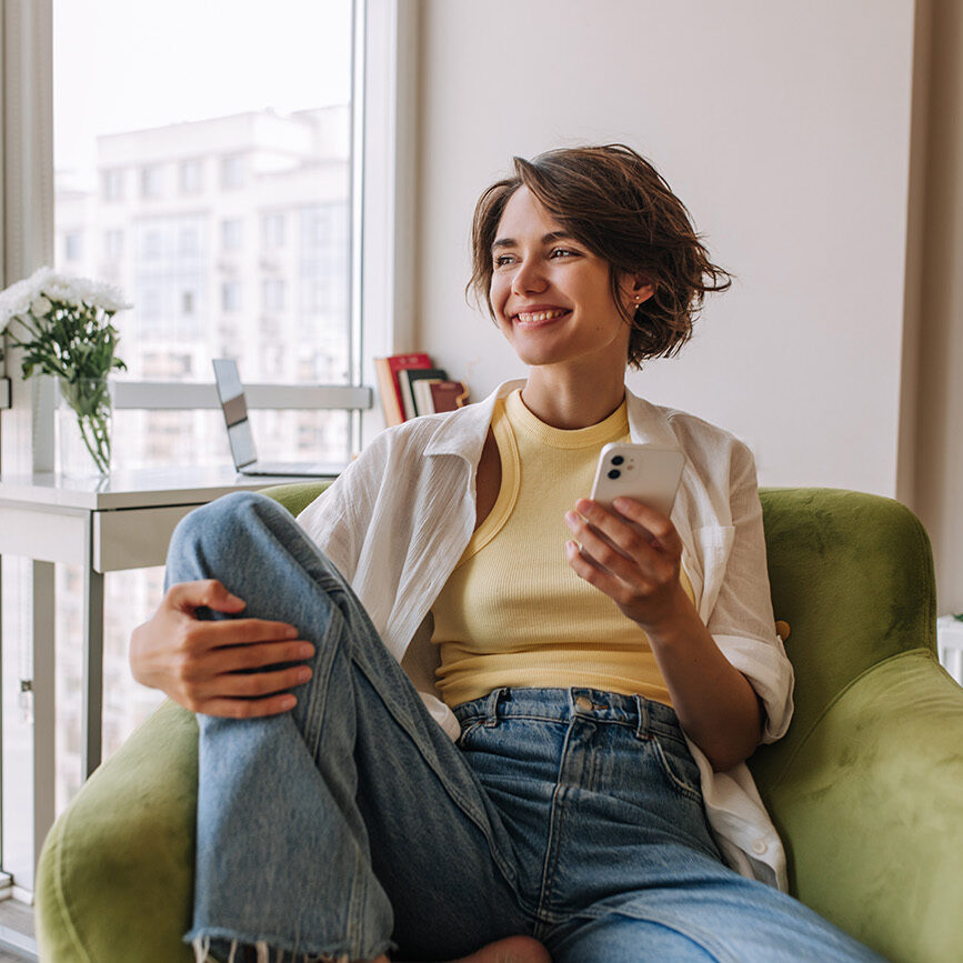 Attractive european woman looking at window . Brunette short hair is sitting on the green chair, smiling holding phone wear white shirt. Concept of lifestyle, use technology